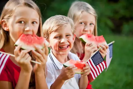 Three happy children celebrate with watermelon and American flags.